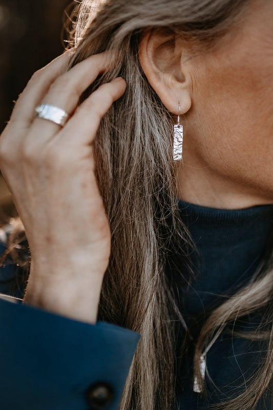 model wearing sterling silver drop earrings with thin rectangle pendant with a captivating driftwood texture