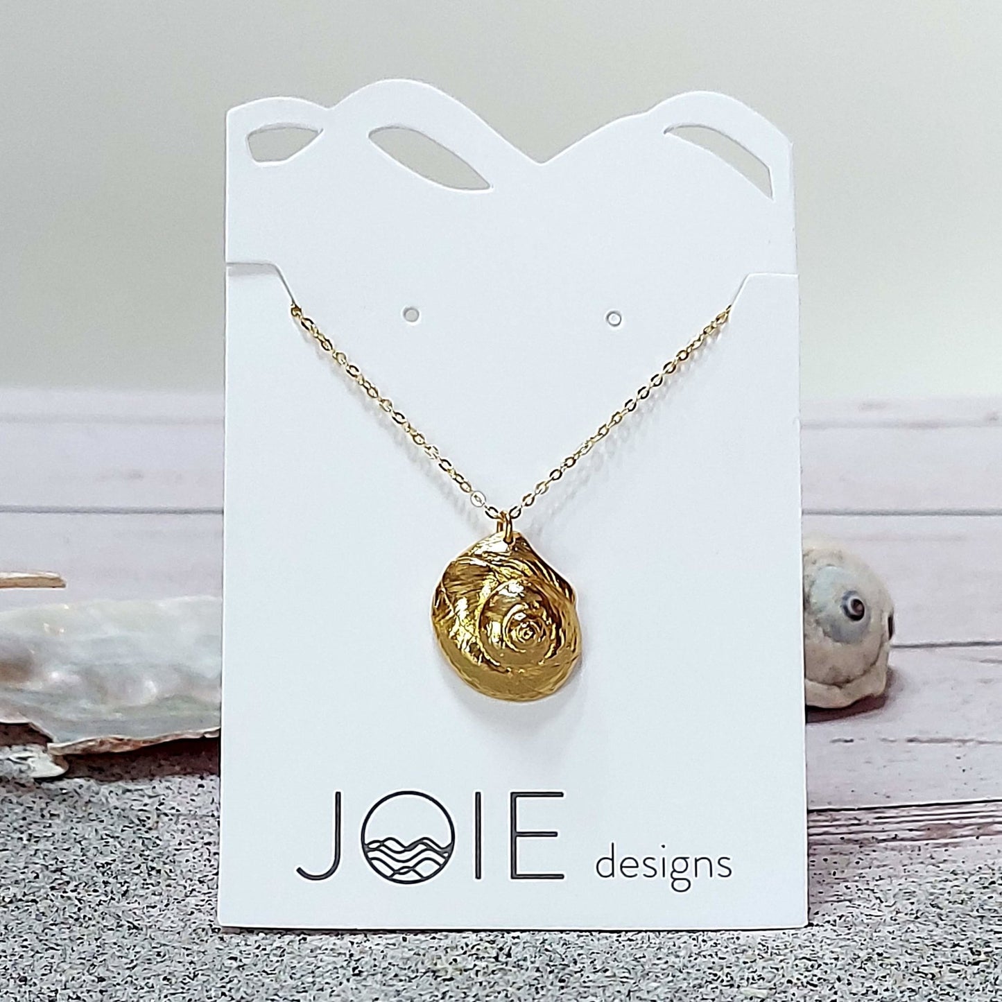 18k plated gold Luna moon snail seashell necklace on a white jewelry card with sand and shells, ocean inspired jewelry