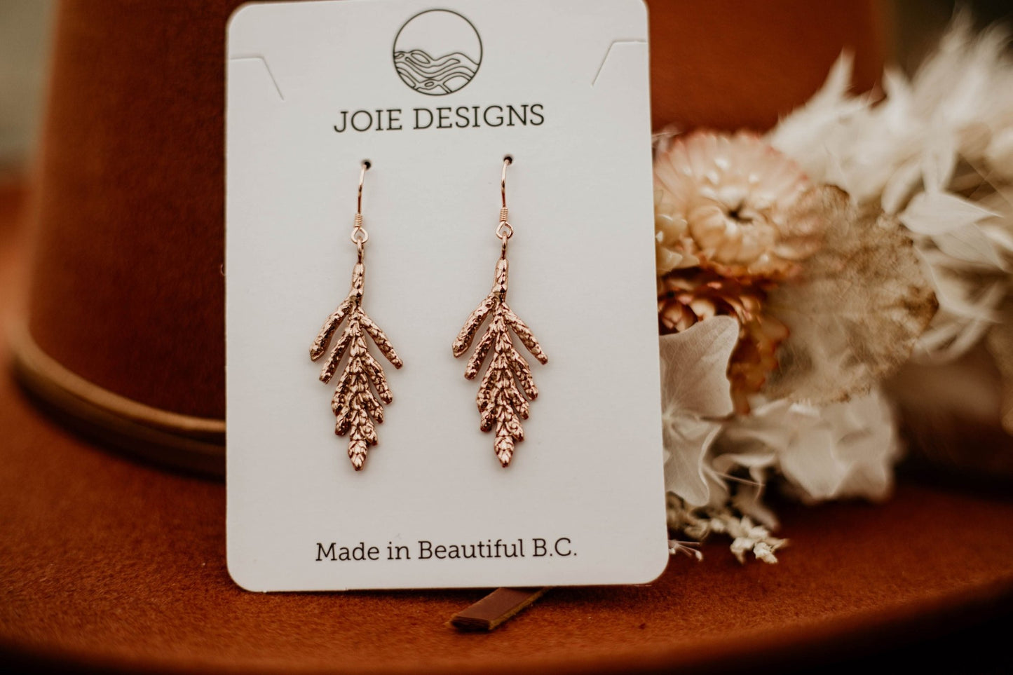 18k rose gold plated cedar leaf design dangle earrings on a jewelry card with dried flowers on wool hat