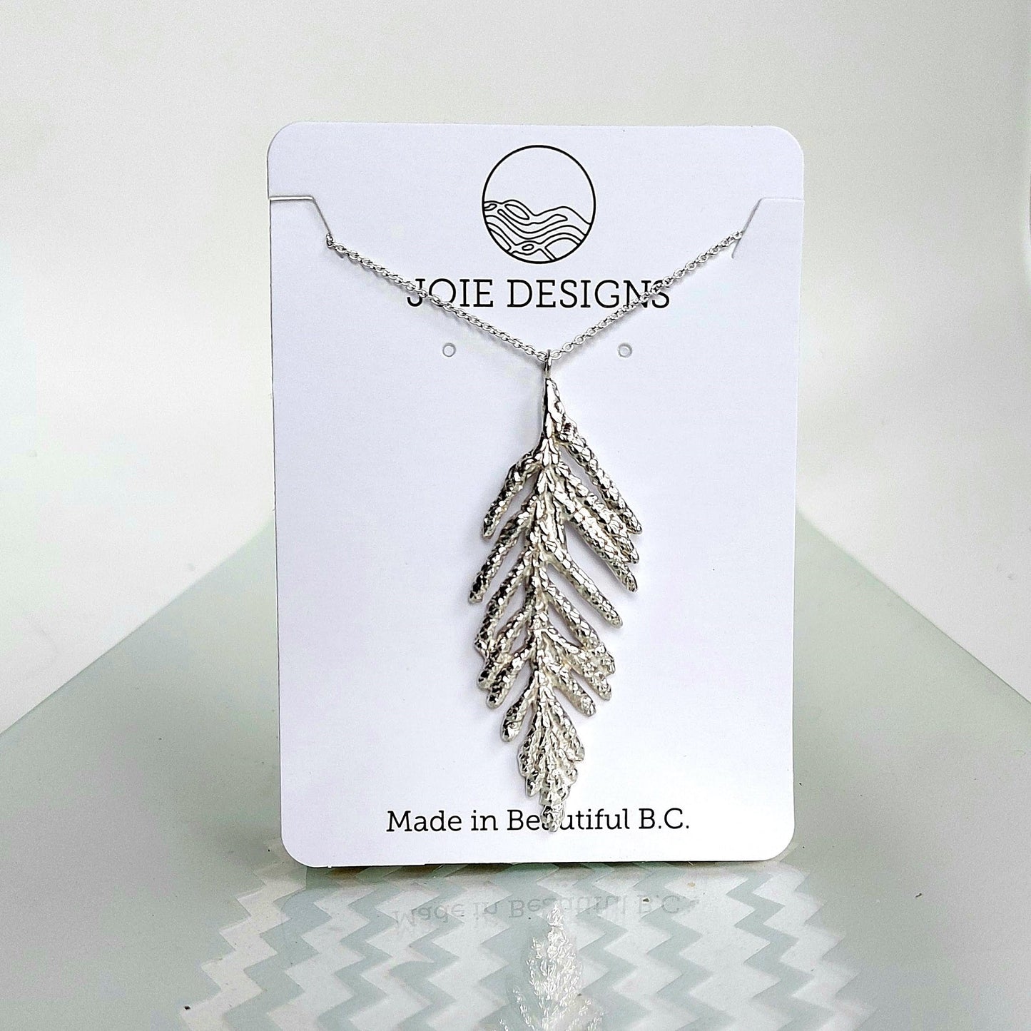 Sterling silver  arborvitae cedar tree leaf pendant on a silver necklace chain shown on a white jewelry card