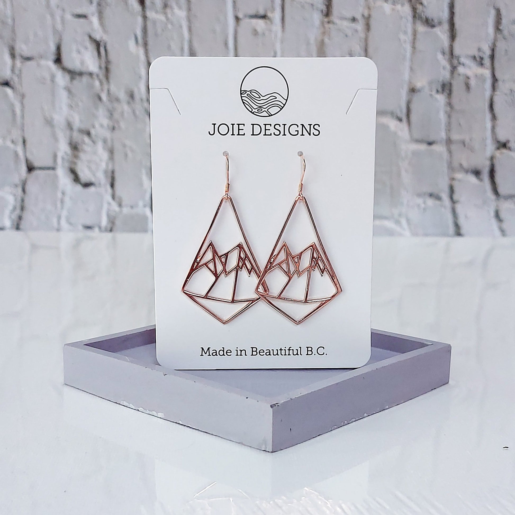 18k rose gold mountain earrings in diamond shape showcased on a jewellery card on purple stand with white brick background