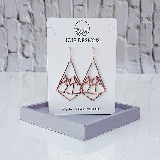 18k rose gold mountain earrings in diamond shape showcased on a jewellery card on purple stand with white brick background