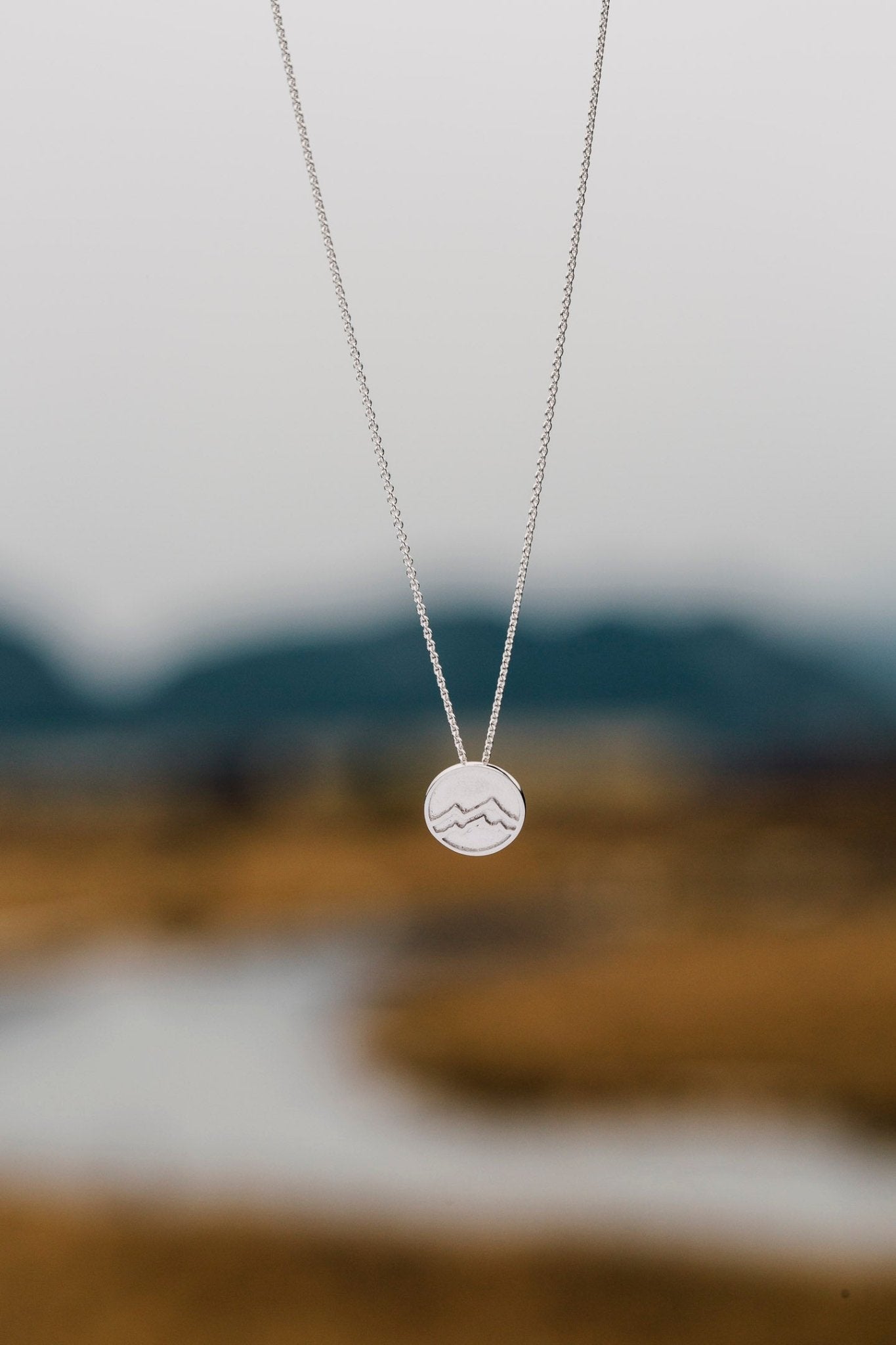 Small Mountain medallion silver pendant necklace shown with blurred blurred river background